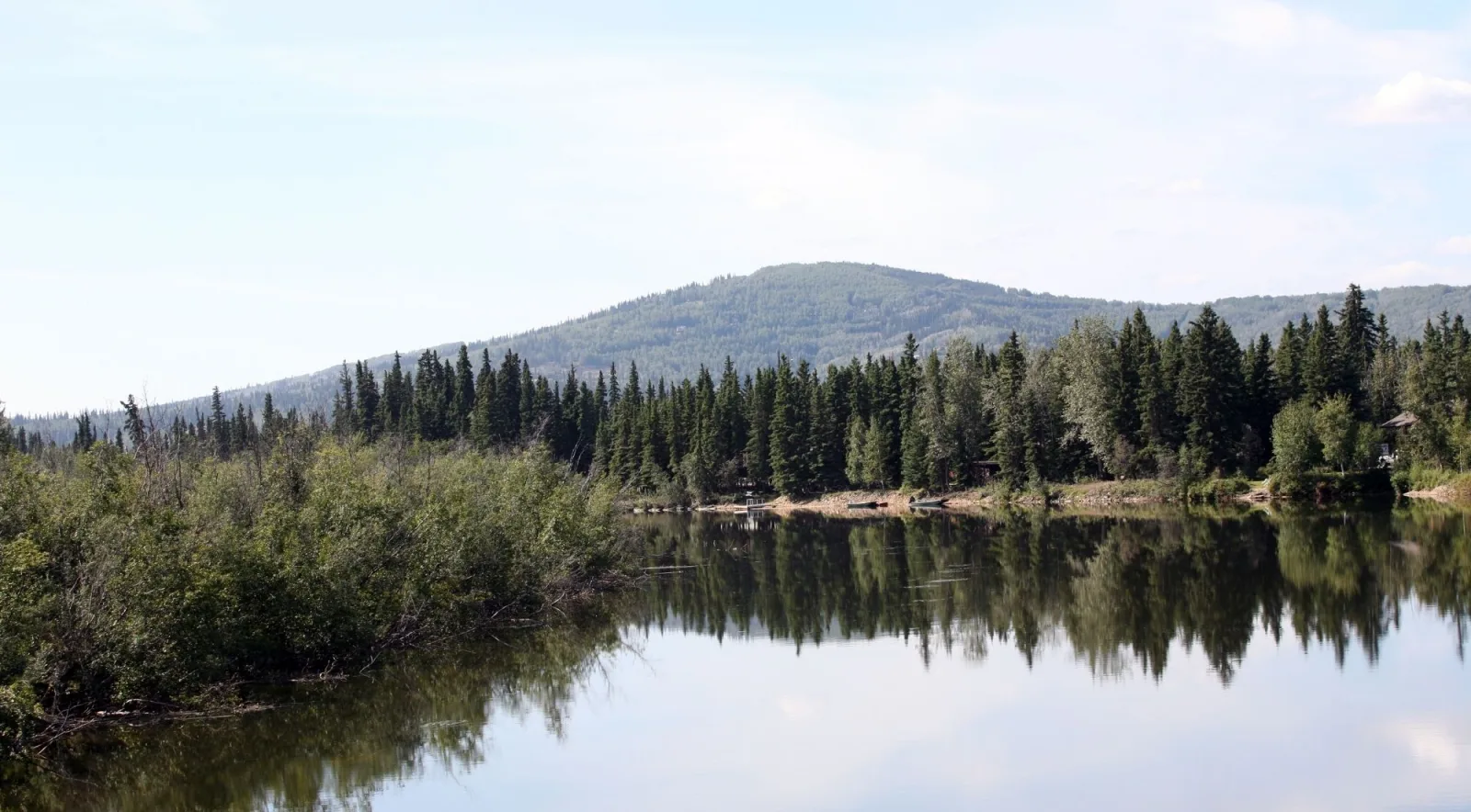 Calm river reflecting green spruce forest and a rounded hill under a pale summer sky